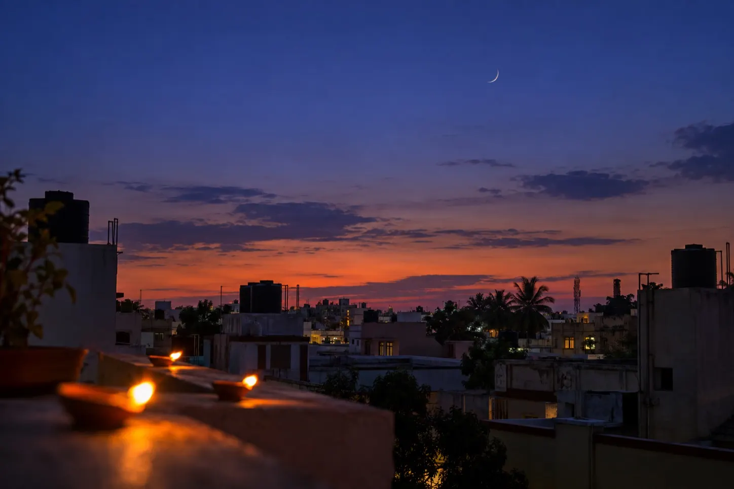 A thin crescent moon high over an Indian rooftop after sunset, oil lamps burning out of focus along the parapet