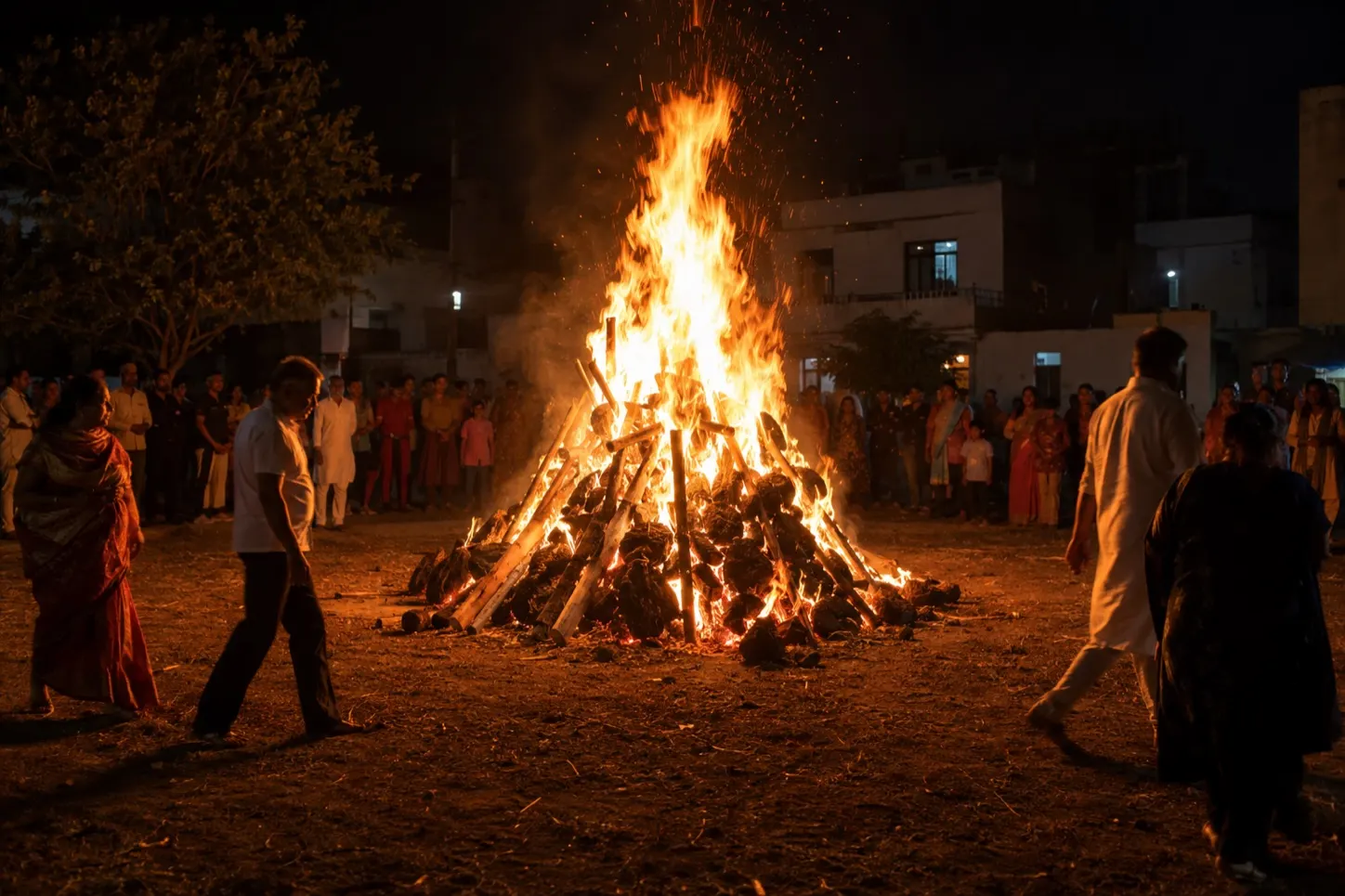 A Holika Dahan bonfire of stacked wood and dung cakes burning at night, a ring of people watching from the edge of the firelight